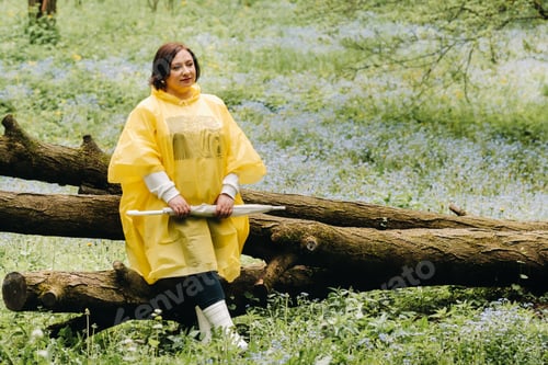 Preview: a woman in a yellow raincoat with an umbrella sits on a fallen tree in the forest in summer