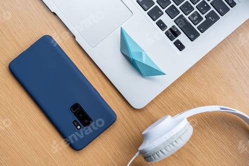 Preview: High angle shot of a blue cellphone, headphones, a laptop, and an origami on a wooden surface