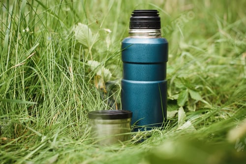 Preview: Thermos and mug standing on the grass outdoors. Steam rising from a steel mug of hot tea. Vacuum