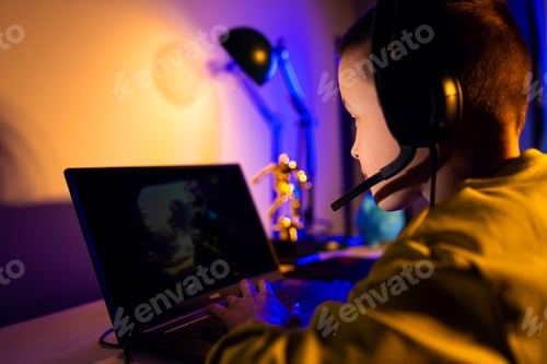 Preview: Young excited boy gamer with headphone playing video game on laptop at home.