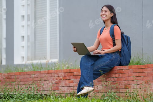 Preview: Happy young asian university student working on her laptop sitting on a brick wall in the campus gar