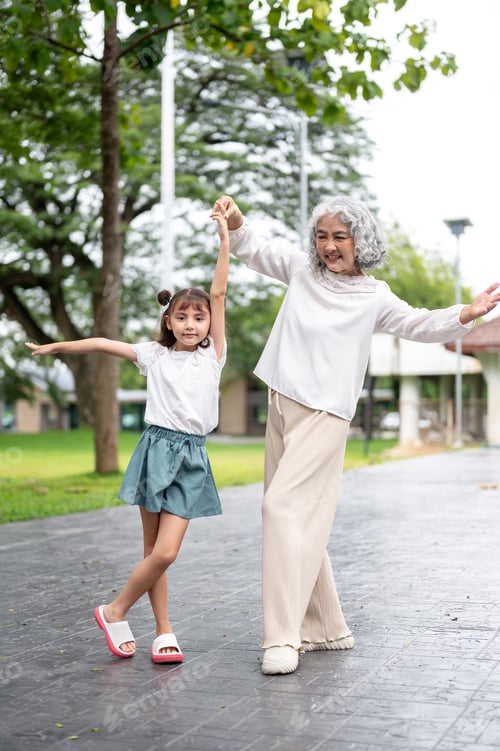 Preview: Cute kid girl granddaughter dancing with old woman grandmother on pavement in the park or garden.