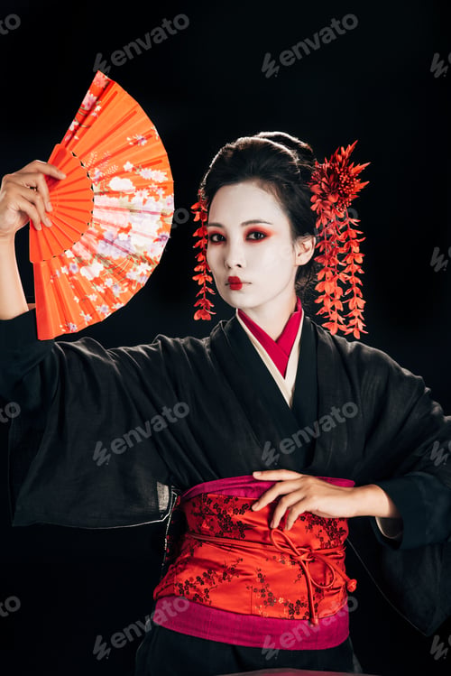 Preview: beautiful geisha in black kimono with red flowers in hair posing with traditional hand fan isolated