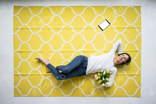 Preview: Smiling woman wearing headphones lying on carpet with bunch of tulips