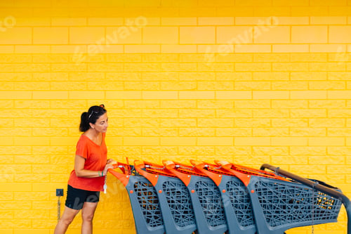 Preview: Woman pushing shopping carts against yellow wall in supermarket parking lot