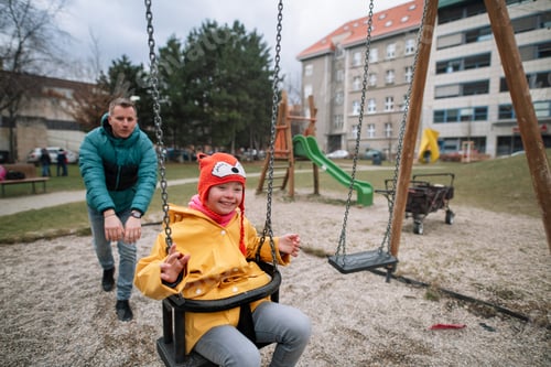 Preview: Father pushing his little daughter with Down syndrome on swing outdoors in playgraound