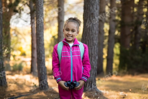 Preview: School-aged girl standing in pine forest wearing pink tracksuit, holding binoculars and backpack
