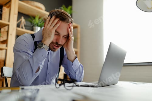 Preview: Male architect in stress holding his head with hands sitting at the table in his office.