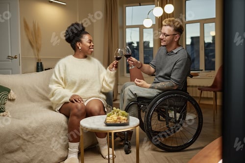 Preview: Cheerful Black Woman Clinking Glasses on Date with Man Using Wheelchair at Home