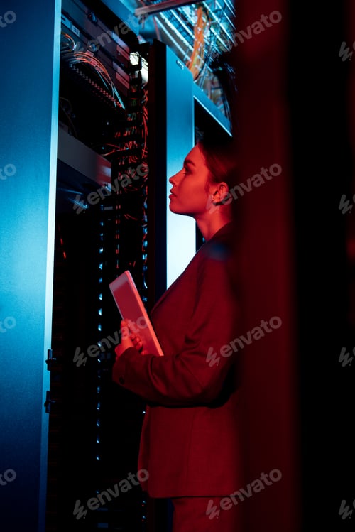 Preview: selective focus of attractive businesswoman looking at server rack and holding digital tablet