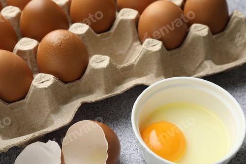 Preview: Many whole raw chicken eggs and broken one in bowl on grey table, closeup