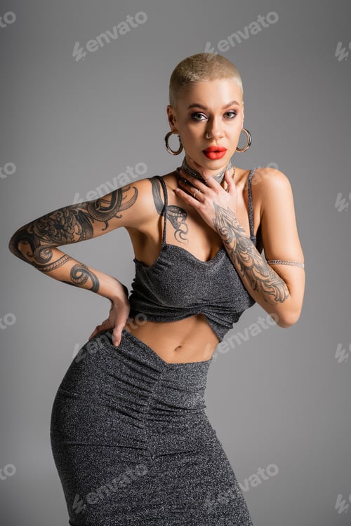 Preview: seductive tattooed woman in lurex skirt and crop top touching necklaces while posing with hand on