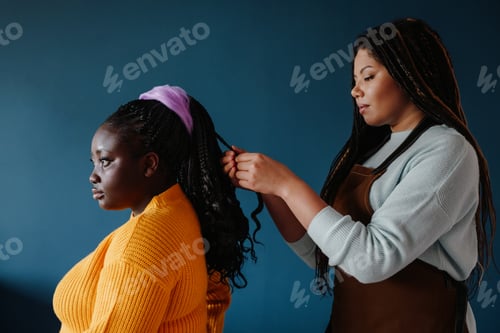 Preview: Side view of confident African hairdresser braiding hair to a female customer on blue background