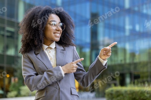 Preview: Confident businesswoman in a suit pointing with both hands outside modern office building in urban