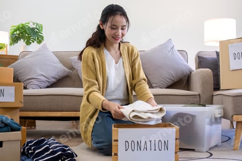 Preview: Cute Asian woman sitting next to sofa in living room sorting unwanted clothes for donation.