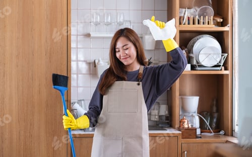 Preview: A woman wearing protective glove, holding a broom and dancing while cleaning in the kitchen at home