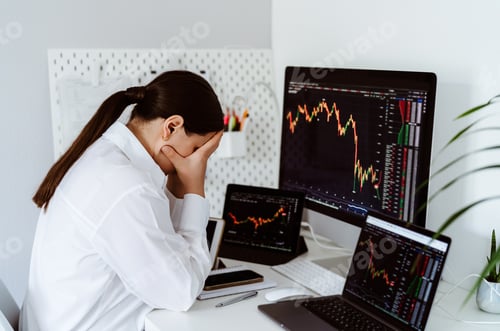 Preview: Young female cryptocurrency trader sitting at desk depressed and upset looking at charts falling