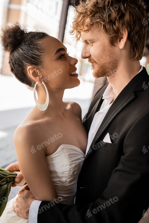 Preview: joyful and elegant interracial newlyweds looking at each other on street, wedding in urban setting