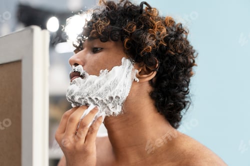 Preview: Handsome young Indian man applies shaving moisturizing foam on face looking at his mirror reflection
