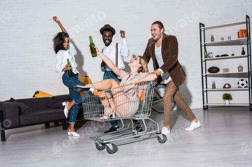 Preview: happy stylish girl riding in shopping cart near multicultural friends holding bottles of beer