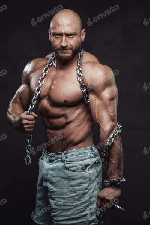Preview: Serious hairless bodybuilder poses with steel chains in dark background
