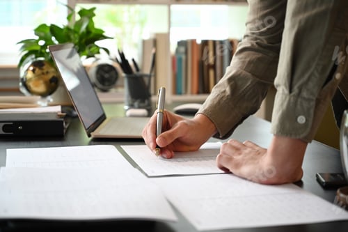 Preview: Cropped shot young man standing at his workplace and writing information on documents.