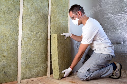Preview: Worker in protective goggles and respirator insulating rock wool insulation in wooden frame