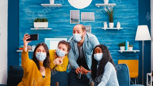 Preview: Multiracial friends sitting on sofa with protection masks, taking selfie