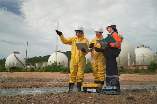 Preview: Workers in protective gear discuss project near industrial storage tanks on a cloudy day