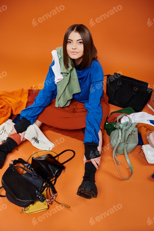 Preview: young woman sitting around handbags and warm clothes on orange backdrop, black friday concept