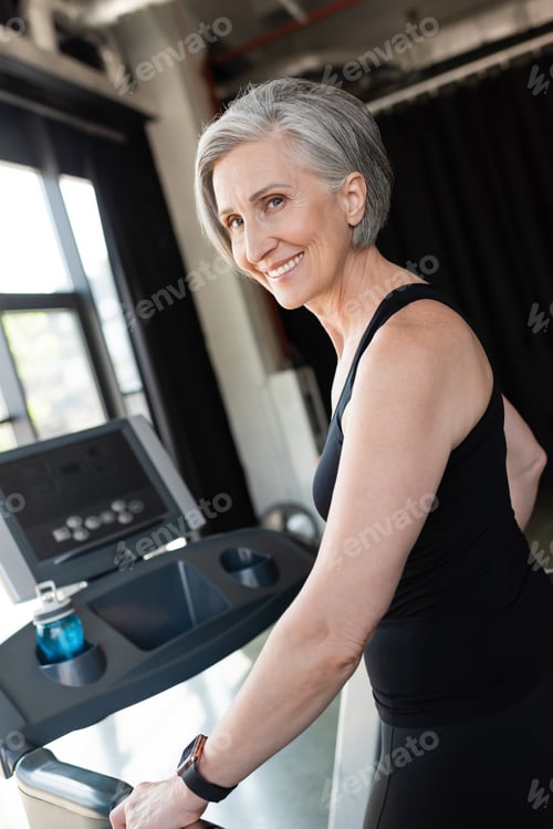 Preview: happy senior woman with grey hair running on treadmill next to sports bottle with water