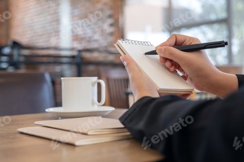 Preview: Closeup image of a hand holding and writing on blank notebook with coffee cup on table in cafe