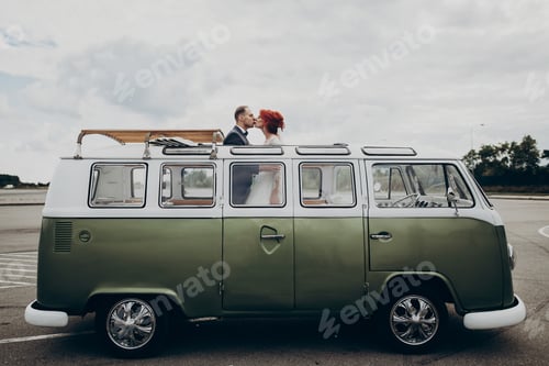 Preview: Bride and stylish groom having fun and laughing, on top of retro car