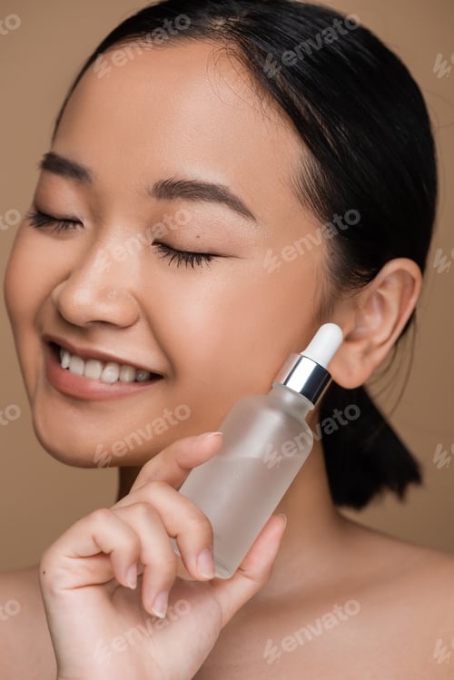 Preview: Close up view of smiling asian model with closed eyes holding serum isolated on brown