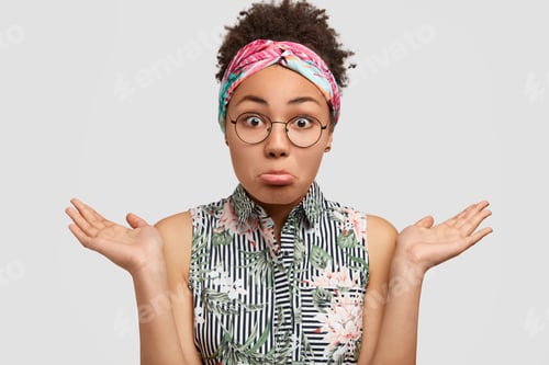 Preview: Beautiful hesitant African American female curves lower lip, shrugs shoulders, wears blouse and head