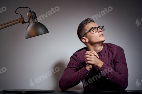 Preview: business man with glasses and a shirt sitting at a table in a dark room with a lamp cropped view