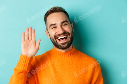 Preview: Close-up of friendly young man smiling, waving hand to say hello, greeting you, standing over light