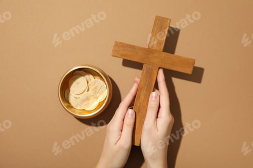 Preview: Wooden cross in hands and liturgical bread on brown background, top view