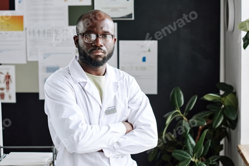 Preview: Portrait of confident African practitioner standing in lacoat with his arms crossed and looking at
