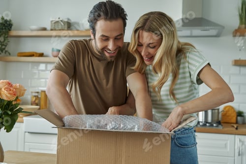 Preview: Happy young couple unpacking box while standing at the domestic kitchen