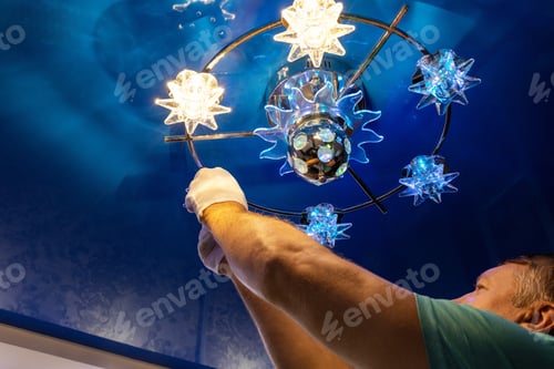 Preview: A man at home changes a light bulb in a chandelier with a blue false ceiling.