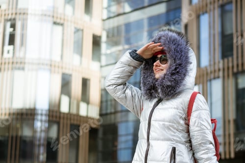 Preview: Happy young woman in sunglasses in a winter jacket with a backpack near the building. Travel concept