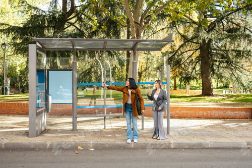Preview: Two young women friends standing at a bus stop, pointing for an incoming bus