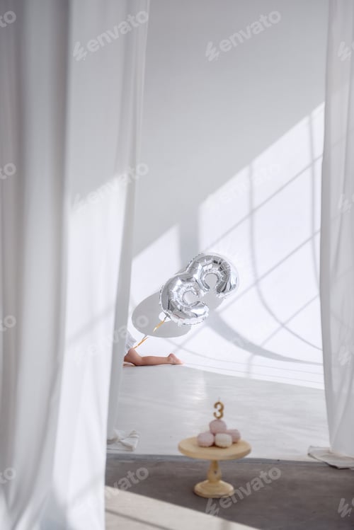 Preview: A little, happy girl in a white dress, barefoot, holding a balloon number three in her hands.