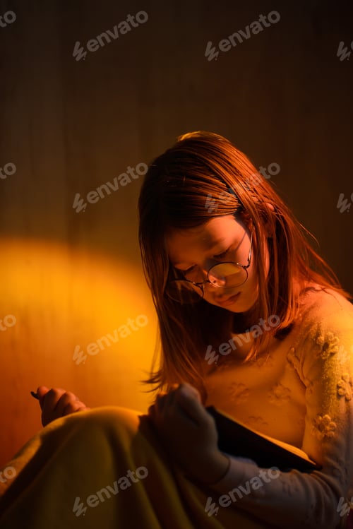 Preview: A girl with loose hair reads a book in the evening by the light of a lamp.