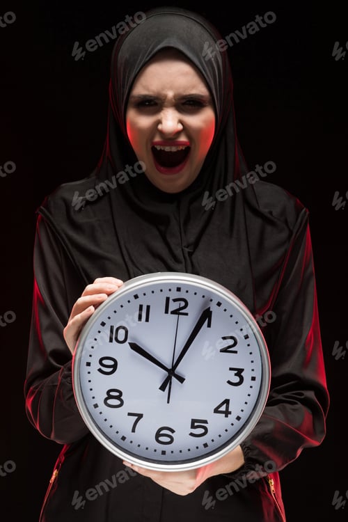 Preview: Portrait of beautiful serious scared frightened young muslim woman wearing black hijab holding clock