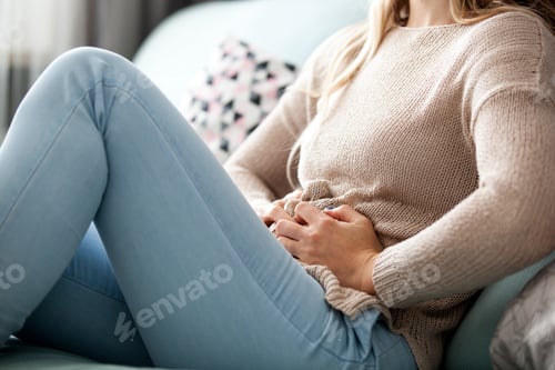 Preview: Young woman suffering from abdominal pain while sitting on sofa at home