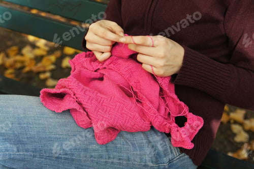 Preview: hands of woman knitting a pink sweater while sitting on park bench in autumn