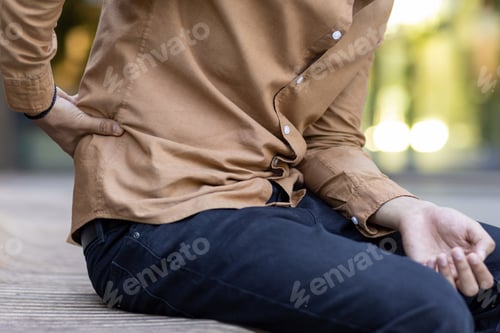 Preview: Person experiencing lower back pain while sitting on wooden bench in outdoor setting, wearing