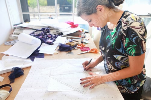 Preview: Focused designer drafting patterns on a worktable surrounded by fabric and tools.